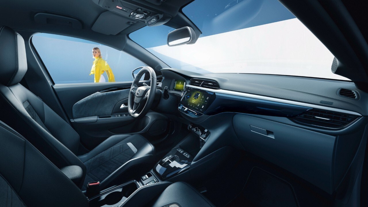 Widok z boku czarnego wnętrza samochodu Opel Corsa Electric od strony siedzenia pasażera, w tle widać kobietę w żółtej koszuli